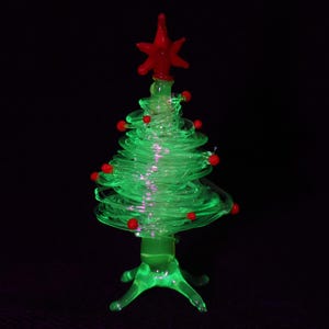 Könnte beinhalten: Eine kleine Weihnachtsbaumfigur aus Glas mit einem gewundenen, durchscheinenden hellgrünen Glaskörper. Der Baum ist mit winzigen, runden roten Glasornamenten verziert und wird von einem markanten, sechsstrahligen roten Glasstern gekrönt.