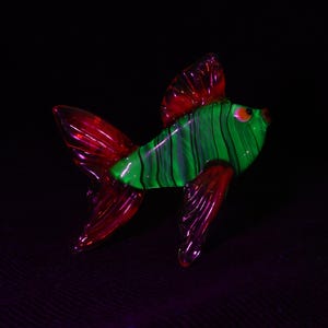 Könnte beinhalten: Eine Glasfisch-Figur mit einem leuchtend grünen Körper und dünnen schwarzen vertikalen Streifen. Der Fisch hat durchscheinende rote Flossen und einen Schwanz sowie ein kleines orangefarbenes Auge. Das Objekt ist vor einem dunklen Hintergrund.