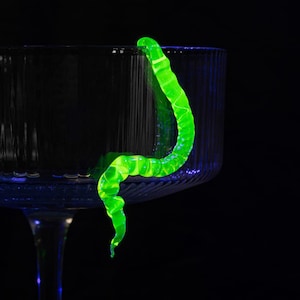 Può includere: Un verme di vetro verde brillante appeso al bordo di una ciotola di vetro trasparente. La ciotola è su uno sfondo nero.