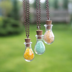 Puede incluir: Tres collares con botellas de vidrio y tapones de corcho. Las botellas están llenas de cristales de diferentes colores. Los collares cuelgan de un fondo verde borroso.