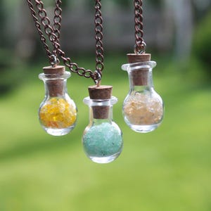 Puede incluir: Tres collares de botella de vidrio con tapones de corcho y cristales de diferentes colores en el interior. Los collares están hechos con cadenas de cobre.