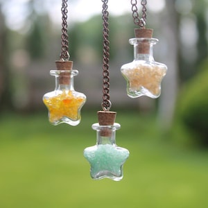 Puede incluir: Tres botellas de vidrio en forma de estrella con tapas de corcho y llenas de purpurina de colores en cadenas plateadas. Las botellas cuelgan sobre un fondo verde borroso.