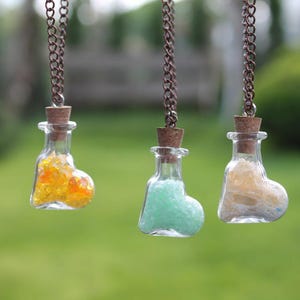 Puede incluir: Tres botellas de vidrio en forma de corazón llenas de arena colorida en cadenas delicadas. Las botellas están suspendidas de las cadenas y cuelgan frente a un fondo verde borroso.