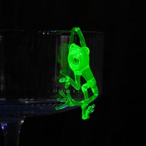 May include: A green glass frog figurine hanging from the rim of a clear glass goblet.