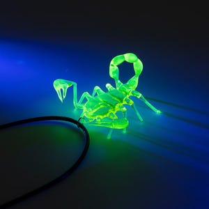 May include: A bright green glass scorpion figurine glows under a black light. The scorpion is posed with its tail raised and its claws extended.