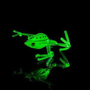 May include: A green glass frog figurine with black spots. The frog is posed in a jumping position with its legs extended. The figurine is illuminated from below, casting a glow on the glass.