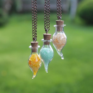 Puede incluir: Tres collares de botella de vidrio con tapones de corcho y cristales de diferentes colores en su interior. Los collares cuelgan de un fondo verde claro.