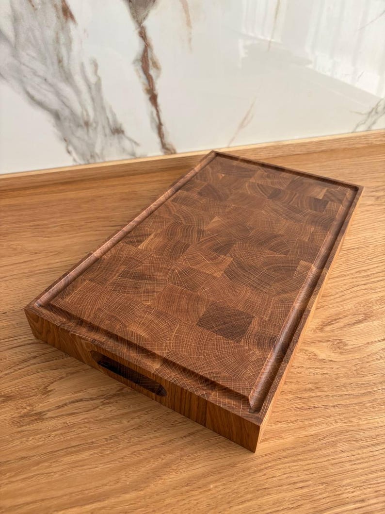 Puede incluir: Tabla de cortar rectangular de madera marr&oacute;n oscuro con veta de extremo. Tiene un canal para jugos y un asa recortada. El patr&oacute;n de la veta de la madera es visible en toda la tabla. Mide aproximadamente 30 cm x 45 cm.