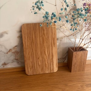 Può includere: Un tagliere rettangolare in legno con angoli arrotondati. La tavola presenta un motivo a venature naturali del legno ed è esposta su una superficie in legno. Un piccolo vaso quadrato in legno contiene fiori blu secchi. Lo sfondo presenta un muro marmorizzato.