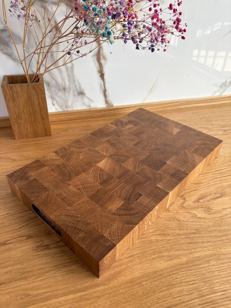 Puede incluir: Una tabla de cortar rectangular de bloque de carnicero de madera marr&oacute;n claro. La tabla tiene un asa recortada en un lado. El patr&oacute;n de la veta de la madera es visible. Un jarr&oacute;n de madera con flores coloridas est&aacute; en el fondo.