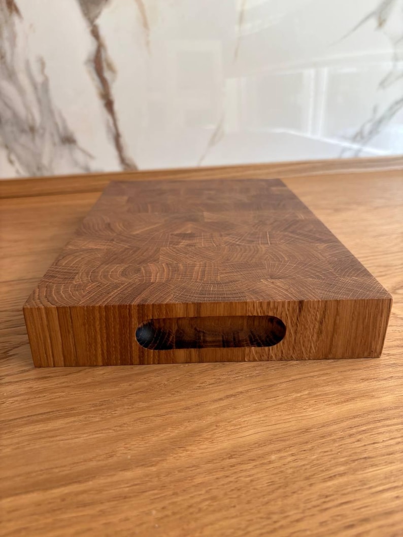 Puede incluir: Una tabla de cortar rectangular de madera marr&oacute;n claro. La tabla tiene un asa incorporada y est&aacute; colocada sobre una superficie de madera. El patr&oacute;n de la veta de la madera es visible en la parte superior y los lados de la tabla.