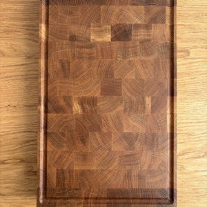 Puede incluir: Una tabla de cortar rectangular de madera con una superficie de testa de color marr&oacute;n oscuro. La tabla tiene un borde empotrado y est&aacute; hecha de varias piezas de madera. El patr&oacute;n de la veta de la madera es visible en toda la tabla.