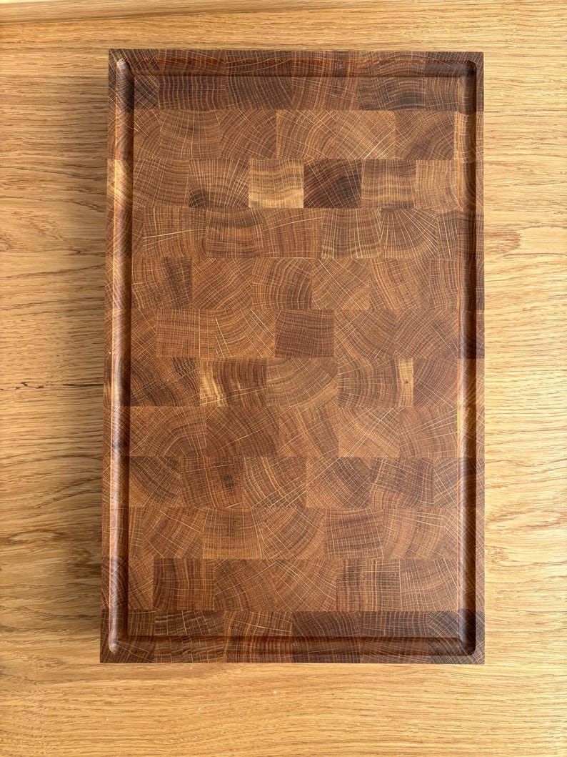 Puede incluir: Una tabla de cortar rectangular de madera con una superficie de testa de color marr&oacute;n oscuro. La tabla tiene un borde empotrado y est&aacute; hecha de varias piezas de madera. El patr&oacute;n de la veta de la madera es visible en toda la tabla.