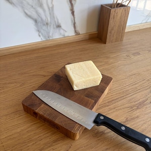 Può includere: Un tagliere in legno con un blocco di formaggio e un coltello in acciaio inossidabile con manico nero. Il tagliere è composto da piccoli pezzi di legno quadrati. Il formaggio è di colore giallo chiaro.
