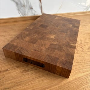 Puede incluir: Una tabla de cortar rectangular de madera de testa hecha de madera marr&oacute;n clara y oscura. La tabla tiene un asa recortada en un lado. El patr&oacute;n de la veta de la madera es visible en toda la tabla.