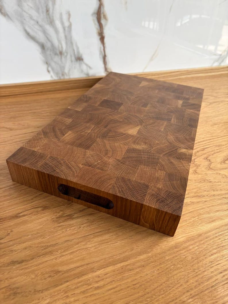 Puede incluir: Una tabla de cortar rectangular de madera de testa hecha de madera marr&oacute;n clara y oscura. La tabla tiene un asa recortada en un lado. El patr&oacute;n de la veta de la madera es visible en toda la tabla.