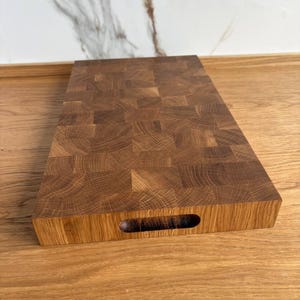 Puede incluir: Una tabla de cortar rectangular de madera clara con un dise&ntilde;o de testa. La tabla tiene un recorte de asa en un lado y est&aacute; colocada sobre una superficie de madera. La construcci&oacute;n de testa es visible, mostrando los patrones naturales de la madera.