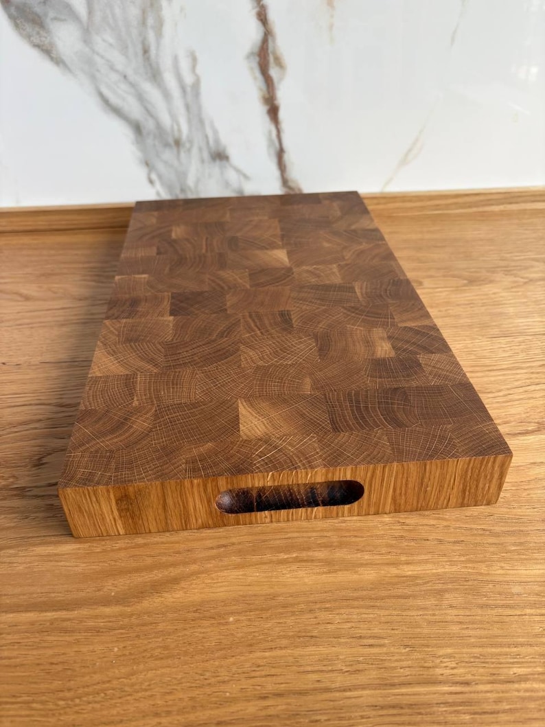 Puede incluir: Una tabla de cortar rectangular de madera clara con un dise&ntilde;o de testa. La tabla tiene un recorte de asa en un lado y est&aacute; colocada sobre una superficie de madera. La construcci&oacute;n de testa es visible, mostrando los patrones naturales de la madera.