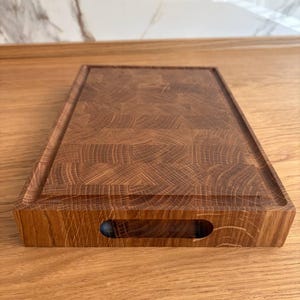 Puede incluir: Tabla de cortar rectangular de madera con acabado marr&oacute;n oscuro. La tabla tiene un canal para jugos alrededor del per&iacute;metro y un recorte para el asa en un lado. La veta de la madera es visible, mostrando la textura natural del material. Mide aprox. 30 cm x 40 cm.