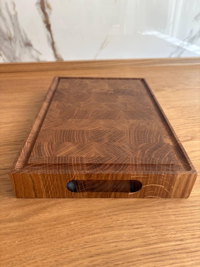Puede incluir: Tabla de cortar rectangular de madera con acabado marr&oacute;n oscuro. La tabla tiene un canal para jugos alrededor del per&iacute;metro y un recorte para el asa en un lado. La veta de la madera es visible, mostrando la textura natural del material. Mide aprox. 30 cm x 40 cm.