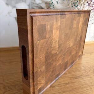 Puede incluir: Tabla de cortar rectangular de madera de testa, de color marr&oacute;n claro y oscuro. La tabla tiene un canal para jugos alrededor del per&iacute;metro y un asa recortada en un lado. El patr&oacute;n de la veta de la madera es visible.