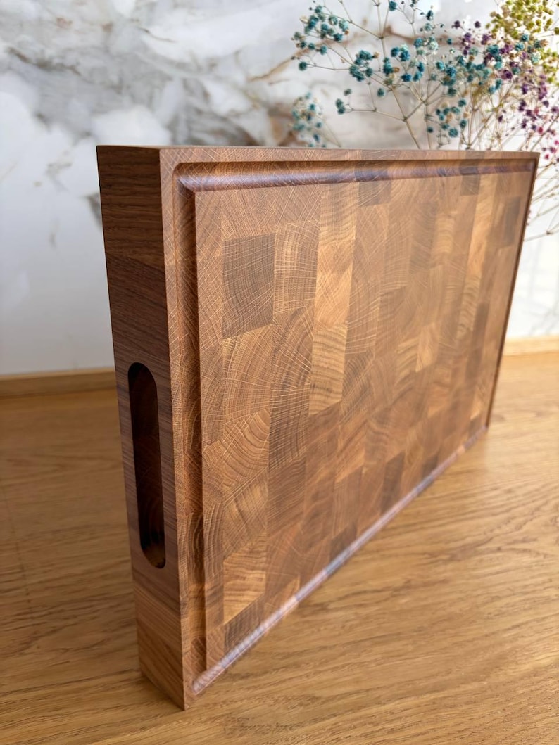 Puede incluir: Tabla de cortar rectangular de madera de testa, de color marr&oacute;n claro y oscuro. La tabla tiene un canal para jugos alrededor del per&iacute;metro y un asa recortada en un lado. El patr&oacute;n de la veta de la madera es visible.