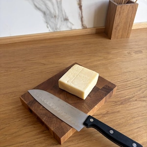 Può includere: Un tagliere in legno con un blocco di formaggio quadrato e un coltello da chef in acciaio inossidabile con manico nero. Il coltello è posizionato diagonalmente sulla tavola. Lo sfondo include un vaso di legno e un muro marmorizzato.