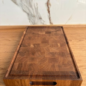 Puede incluir: Tabla de cortar rectangular de madera con superficie de testa marr&oacute;n oscuro. La tabla tiene un canal para jugos empotrado y un recorte para el asa. El patr&oacute;n de vetas de la madera es visible en toda la tabla.