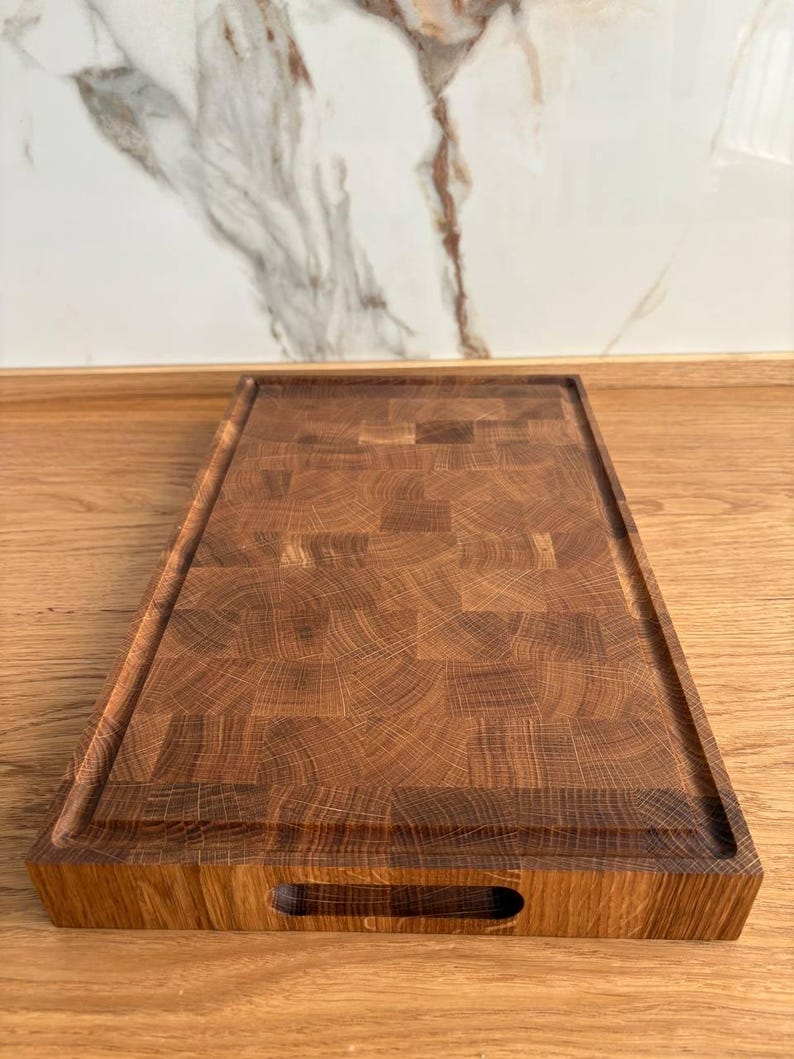 Puede incluir: Tabla de cortar rectangular de madera con superficie de testa marr&oacute;n oscuro. La tabla tiene un canal para jugos empotrado y un recorte para el asa. El patr&oacute;n de vetas de la madera es visible en toda la tabla.