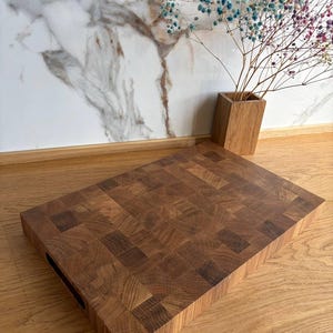 Puede incluir: Una tabla de cortar rectangular de madera marr&oacute;n oscuro. La tabla tiene un asa recortada en un lado. Un jarr&oacute;n de madera con flores secas de colores est&aacute; en el fondo.