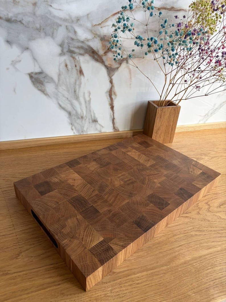 Puede incluir: Una tabla de cortar rectangular de madera marr&oacute;n oscuro. La tabla tiene un asa recortada en un lado. Un jarr&oacute;n de madera con flores secas de colores est&aacute; en el fondo.
