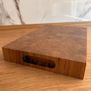 Puede incluir: Una tabla de cortar rectangular de madera con un asa cortada en el lateral. La tabla est&aacute; hecha de madera de testa, mostrando un patr&oacute;n de grano circular. La madera tiene un color marr&oacute;n c&aacute;lido y un acabado liso. La tabla de cortar est&aacute; dise&ntilde;ada para uso en la cocina.