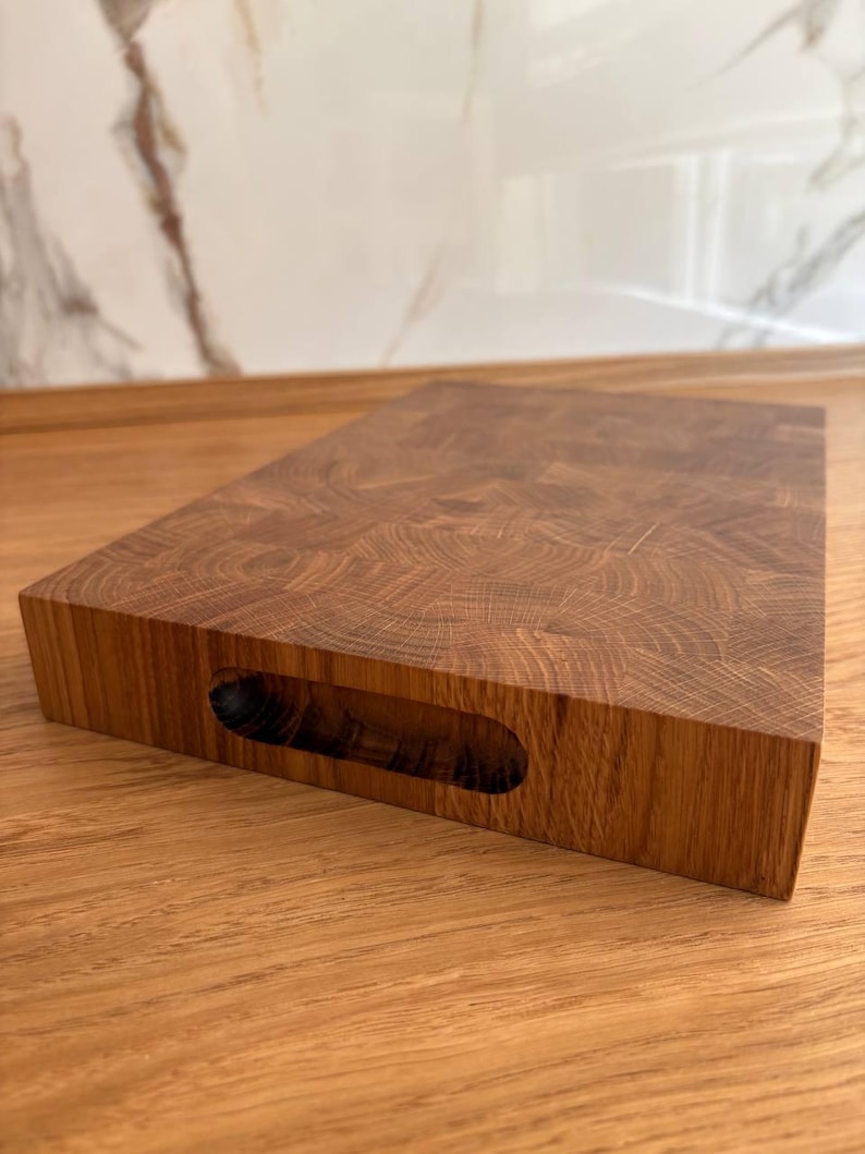 Puede incluir: Una tabla de cortar rectangular de madera con un asa cortada en el lateral. La tabla est&aacute; hecha de madera de testa, mostrando un patr&oacute;n de grano circular. La madera tiene un color marr&oacute;n c&aacute;lido y un acabado liso. La tabla de cortar est&aacute; dise&ntilde;ada para uso en la cocina.