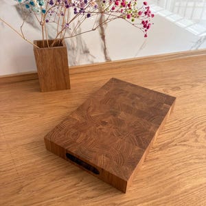 Puede incluir: Tabla de cortar rectangular de bloque de carnicero de madera marr&oacute;n. La tabla tiene un asa oscura y rectangular recortada en un lado. La tabla est&aacute; sobre una superficie de madera con un jarr&oacute;n de flores coloridas en el fondo.