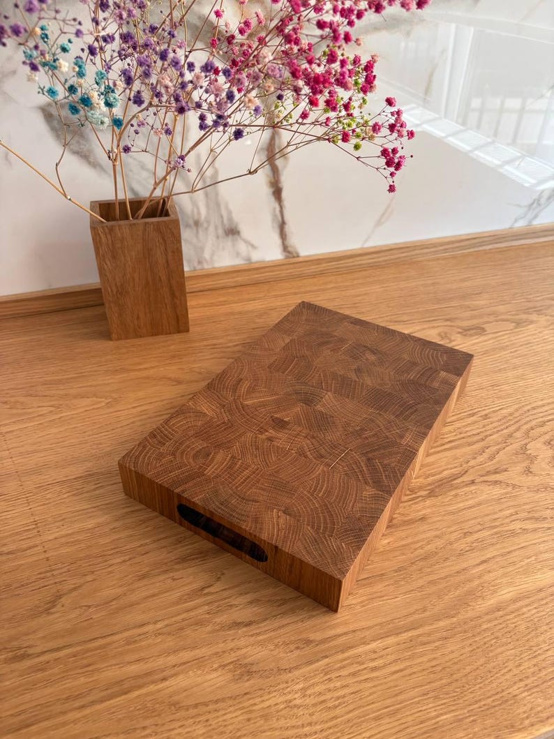 Puede incluir: Tabla de cortar rectangular de bloque de carnicero de madera marr&oacute;n. La tabla tiene un asa oscura y rectangular recortada en un lado. La tabla est&aacute; sobre una superficie de madera con un jarr&oacute;n de flores coloridas en el fondo.
