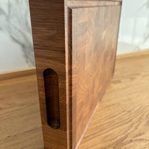 Puede incluir: Tabla de cortar rectangular de madera clara con un hueco para el asa en el lateral. La superficie es texturizada y los bordes ligeramente elevados. Se apoya sobre una superficie de madera. Mide unos 30 cm de largo.