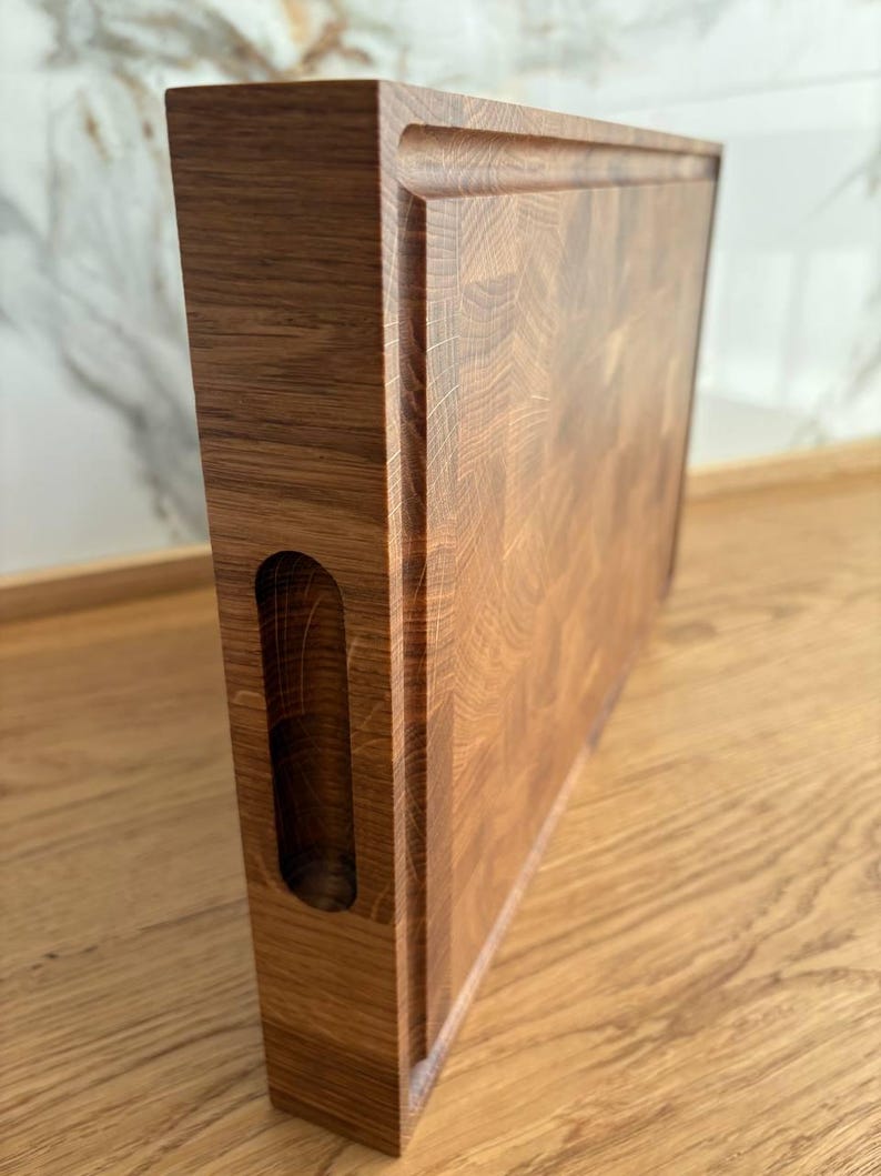 Puede incluir: Tabla de cortar rectangular de madera clara con un hueco para el asa en el lateral. La superficie es texturizada y los bordes ligeramente elevados. Se apoya sobre una superficie de madera. Mide unos 30 cm de largo.