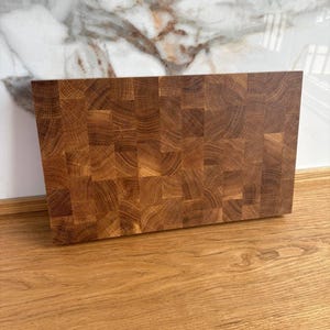 Puede incluir: Una tabla de cortar rectangular de bloque de carnicero de madera marr&oacute;n claro. La tabla est&aacute; construida con peque&ntilde;os trozos cuadrados de madera, creando una superficie texturizada. La tabla est&aacute; sobre una superficie de madera.