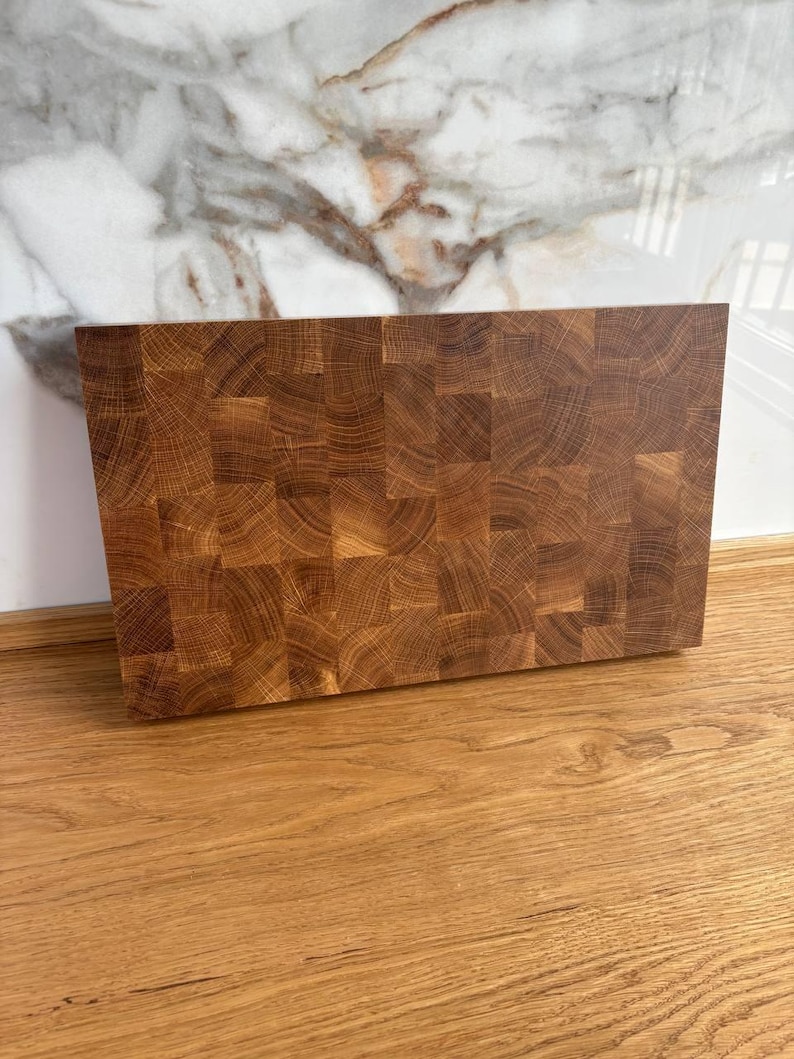 Puede incluir: Una tabla de cortar rectangular de bloque de carnicero de madera marr&oacute;n claro. La tabla est&aacute; construida con peque&ntilde;os trozos cuadrados de madera, creando una superficie texturizada. La tabla est&aacute; sobre una superficie de madera.
