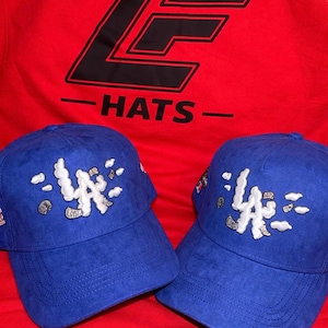 May include: Two blue baseball caps with the letters "LA" in white and gray embroidery. The caps are displayed on a red background, with a red shirt that has the text "HATS" and a logo.