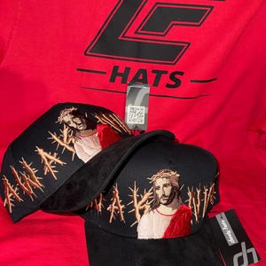 May include: Black baseball caps with embroidered image of Jesus wearing a crown of thorns and a red robe. The word "AMEN" is embroidered in gold on the side. A red shirt with the text "LF HATS" is in the background.