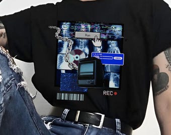 May include: A black t-shirt featuring a graphic print of a computer screen with a blue and white interface. The graphic includes the text "Run" and "REC". The person wearing the shirt is also wearing blue jeans with rips and black fishnet tights.