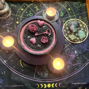 Peut inclure: Scène mystique avec un tapis astrologique noir et violet orné de bougies, de cristaux et d'objets décoratifs. Un plateau circulaire central contient des décorations en forme de cœur et de rose, ainsi qu'une clé. Une statue de serpent argentée et une boîte en bois sont également présentes.