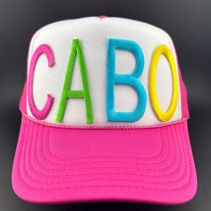 May include: A vibrant pink trucker hat with the word "CABO" embroidered in colourful letters. The hat has a white front panel, a pink brim, and a mesh back. The letters are pink, green, blue, and yellow.