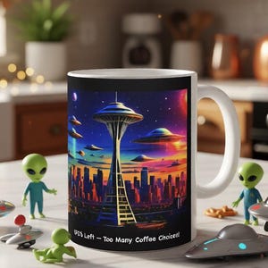 May include: White ceramic coffee mug featuring a colorful illustration of the Seattle Space Needle, UFOs, and a cityscape under a starry sky. The mug has the text "UFO's Left - Too Many Coffee Choices!" surrounded by alien figurines and UFO toys.