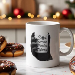 May include: White coffee mug with the text "Coffee Because Murder Wrong" held by a black hand graphic. The mug is surrounded by chocolate frosted donuts with colorful sprinkles and a Christmas tree in the background.