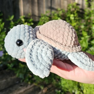 May include: A handmade plush turtle with a light blue head and flippers, a tan shell, and a black eye. The turtle is made of soft, textured yarn and is a cute, cuddly toy.
