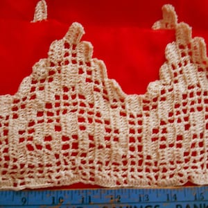 May include: Close-up of a cream-colored crocheted lace trim with a scalloped edge. The lace is against a red background and a blue ruler is visible at the bottom of the image.