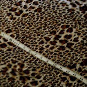 May include: Close-up of a leopard print fabric with a measuring tape. The fabric features a pattern of brown spots on a cream background. The measuring tape shows measurements in inches.