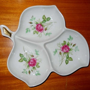 May include: A white ceramic serving dish shaped like a leaf with three sections. Each section has a pink rose and green leaves painted on it. The dish has a gold handle.